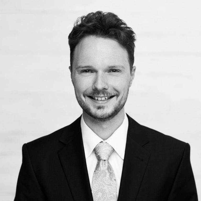Jörn Müller Mann in Anzug mit Krawatte, lächelt vor einem hellen, neutralen Hintergrund.